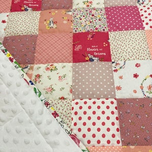 May include: A patchwork quilt with a variety of floral and polka dot patterns in shades of pink, white, and beige. The quilt features the text "Made of Flowers and Dreams" in a pink font.