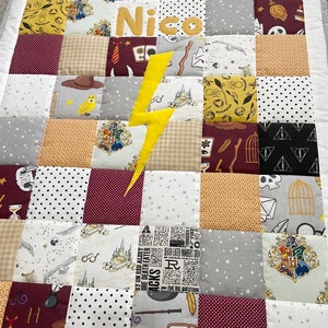 Könnte beinhalten: Eine mehrfarbige Patchwork-Decke mit Harry-Potter-Motiv. Die Decke zeigt verschiedene Harry-Potter-Symbole, darunter das Hogwarts-Wappen, das Symbol der Heiligtümer des Todes und einen Blitz. Die Decke enthält auch einen Namen, "Nico", in gelben Buchstaben.