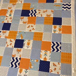 May include: A patchwork baby quilt with a variety of patterns in blue, orange, and white. The quilt features foxes, arrows, stars, and geometric patterns.