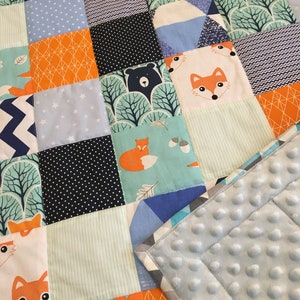 May include: A patchwork baby quilt with a variety of blue, green, orange, and white fabrics. The quilt features a fox, bear, and geometric patterns. The back of the quilt is a soft, light blue fabric with a textured surface.