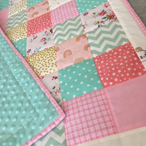 Könnte beinhalten: Eine Patchwork-Babydecke mit einer Vielzahl von rosa, grünen und weißen Stoffen. Die Decke besteht aus Quadraten mit verschiedenen Mustern, darunter Punkte, Sterne und Regenbögen. Die Decke ist mit einem rosa-weißen Besatz eingefasst.