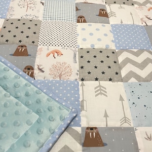 Könnte beinhalten: Eine Babydecke mit Patchwork-Design, das verschiedene Tierdrucke zeigt, darunter Walrosse, Füchse und Hirsche. Die Decke besteht aus weichen, blauen und grauen Stoffen mit weißen Punkten und Sternen. Die Decke hat eine hellblaue Minky-Rückseite.