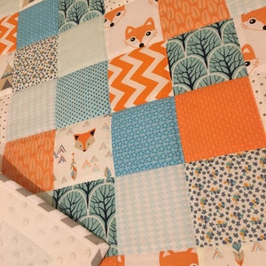 May include: A patchwork baby quilt with a woodland theme. The quilt features a variety of fabrics in shades of orange, blue, and white, with patterns including foxes, trees, and geometric shapes.