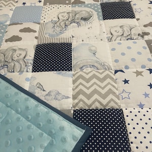 May include: A patchwork quilt with squares featuring elephant illustrations, stars, clouds, and polka dots in blue, grey, and white. The quilt has a soft, light blue, textured border.