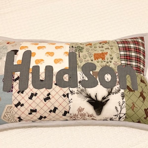 May include: A patchwork pillow with a grey felt name "Hudson" in the centre. The pillow is made of various fabrics with different patterns, including plaid, floral, and animal prints.