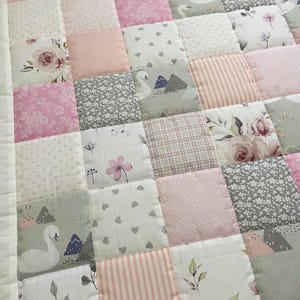 May include: A patchwork quilt with a variety of patterns and colours. The quilt features floral, swan, and heart designs in pink, grey, and white. The quilt is bordered with white fabric.