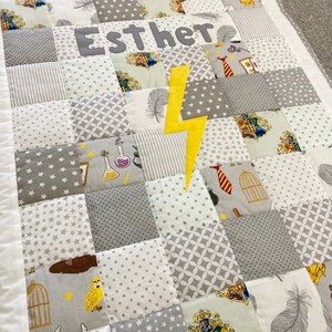 May include: A gray and white patchwork baby quilt with a yellow lightning bolt and the name "Esther" embroidered in gray. The quilt features various Harry Potter themed patterns, including the Hogwarts crest, a feather, a potion bottle, and a birdcage.