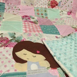 Könnte beinhalten: Eine Patchwork-Decke mit einer hellblauen Minky-Rückseite. Die Decke besteht aus verschiedenen floralen und geometrischen Mustern in Rosa, Grün und Weiß. Ein braunes Mädchen mit geschlossenen Augen ist auf die Decke appliziert.