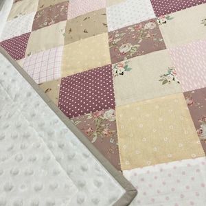 Może przedstawiać: Mata do zabawy dla niemowląt w patchworku z różnymi wzorami kwiatowymi i geometrycznymi w odcieniach różowego, beżowego i brązowego. Mata do zabawy jest wykonana z miękkiej, pikowanej tkaniny i ma białe, wyściełane podłoże.