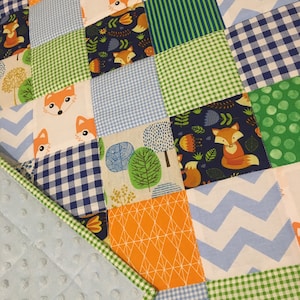 May include: A patchwork baby quilt with a blue and white checkered pattern, green and white checkered pattern, orange and white geometric pattern, and a blue and white chevron pattern. The quilt also features a variety of fox prints and a green and white tree print.