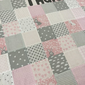 Könnte beinhalten: Eine rosafarbene und graue Patchwork-Babydecke mit dem Namen "Thaleia", der in Schwarz gestickt ist. Die Decke zeigt verschiedene Muster, darunter Herzen, Wolken und Hasen.