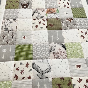 May include: A patchwork quilt featuring a woodland theme. The quilt is made from a variety of fabrics with different patterns, including animals, trees, stars and arrows. The quilt is in shades of green, grey and white.