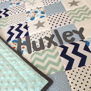 May include: A blue and white patchwork baby quilt with a grey felt name "Huxley" sewn on. The quilt is made of various fabrics with different patterns, including stars, polka dots, and zigzags. The quilt is backed with a soft, light blue minky fabric.
