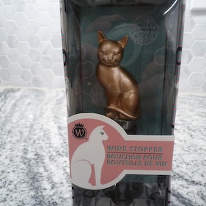 May include: A gold cat-shaped wine stopper in a clear plastic box with a pink and white label that reads "Wine Stopper Bouchon Pour Bouteille de Vin".