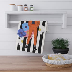 Puede incluir: Un paño de cocina decorativo con un fondo a rayas blancas y negras. El paño presenta una ilustración estilizada de una persona con un traje naranja y zapatos negros, con flores azules y blancas. El paño está colgado de un estante blanco.