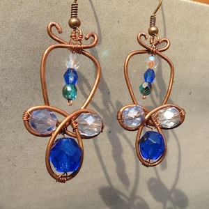 以下が含まれることがあります： 青と透明なガラスビーズが付いた銅線製のイヤリングのペア。イヤリングは涙の形をしていて、上部に吊り下げ用の小さなループが付いています。