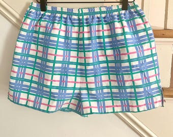 May include: Shorts in a blue, green, pink, and white plaid pattern. The garment is suspended from a wooden hanger. The shorts feature an elasticated waistband and a loose fit. The plaid design is formed by intersecting lines of various colours.