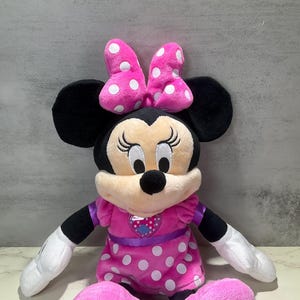 May include: A plush Minnie Mouse doll. The doll has black ears, a pink bow with white polka dots, and a pink dress with white polka dots. The doll's arms and legs are black and white. The doll has a smiling face.