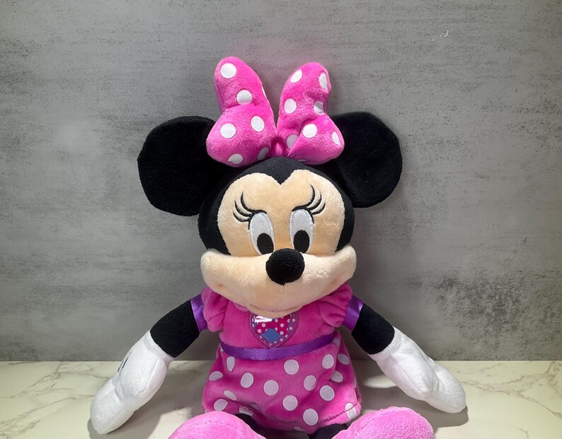 May include: A plush Minnie Mouse doll. The doll has black ears, a pink bow with white polka dots, and a pink dress with white polka dots. The doll's arms and legs are black and white. The doll has a smiling face.