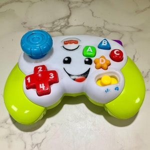 May include: A colorful Fisher-Price toy controller with a smiling face, buttons, and a joystick. The controller features a red cross-shaped button with numbers, and green, blue, and red buttons with letters. The controller has a yellow and green color scheme.