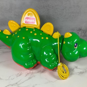 May include: A green and yellow Playskool dinosaur pull toy. The toy has yellow accents, including the fins and a handle. The dinosaur has a yellow pull string and the word "Playskool" is visible on the back.