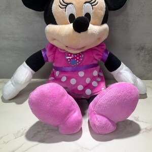 May include: A plush Minnie Mouse doll, featuring a pink dress with white polka dots, a purple waist sash, and large pink feet. The doll has black ears, a black nose, and white gloves.