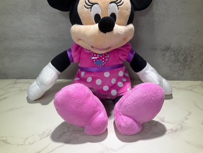 May include: A plush Minnie Mouse doll, featuring a pink dress with white polka dots, a purple waist sash, and large pink feet. The doll has black ears, a black nose, and white gloves.
