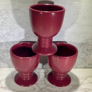 May include: Three burgundy-colored ceramic egg cups. Each cup has a rounded bowl shape, a pedestal base, and a short stem. The cups are arranged in a triangular formation, with one cup stacked on top of the other two.