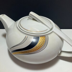 May include: White ceramic teapot with a curved handle and spout. The teapot features a black and gold design on the side, with a matching lid. The teapot is a classic design for serving tea.