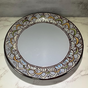 May include: A round decorative plate with a light blue center and a detailed border. The border features a repeating pattern in brown, yellow, green, and blue. The plate has a dark brown rim and is set against a marble-like surface.
