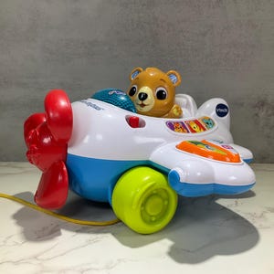 May include: A colorful toy airplane with a smiling bear pilot. The plane is white, blue, and green, with a red propeller and a yellow pull string. It has buttons and illustrations on the wings. The toy is made by VTech.