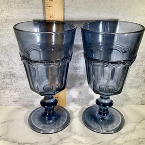 May include: Two blue-tinted vintage water goblets. Each goblet features a faceted bowl, a decorative band, and a round base. The goblets are approximately 7 inches tall.
