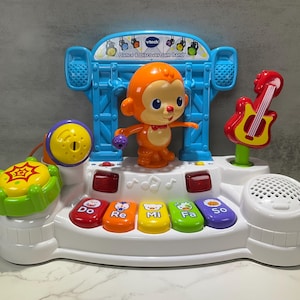 May include: A white and blue VTech "Dance & Discover Jam Band" toy. It features an orange monkey character, a red guitar, and colorful piano keys labeled "Do, Re, Mi, Fa, So." The toy includes a yellow drum and a speaker.