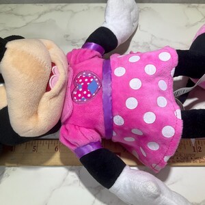 May include: A plush Minnie Mouse doll, lying on a surface. The doll features a pink dress with white polka dots, a heart-shaped patch, and black ears. The doll's face is visible, with black eyes and a black nose.