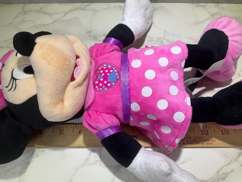 May include: A plush Minnie Mouse doll, lying on a surface. The doll features a pink dress with white polka dots, a heart-shaped patch, and black ears. The doll's face is visible, with black eyes and a black nose.