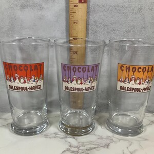 May include: Three clear glass tumblers with a vintage design. Each glass features a rectangular label with the word "CHOCOLAT" in orange, purple, and yellow, above a line of cartoon figures. The text "DELESPAUL-HAVEZ" is below the figures.