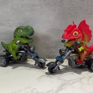 May include: Two toy dinosaur figures riding motorcycles. One dinosaur is green, the other is red and yellow. The motorcycles are silver and black with blue license plates. The toys are on a white surface.