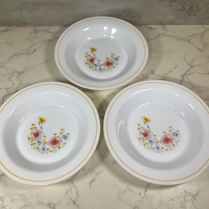 May include: Three white ceramic bowls with a floral design in the center. The flowers are pink, yellow, and blue. Each bowl has a yellow rim. The bowls are arranged on a white and gray surface.