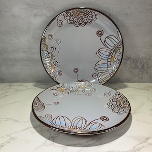 May include: Two ceramic plates with a floral design. The plates are light gray with a brown rim and feature a hand-painted floral pattern in brown, blue, and yellow. The plates are of different sizes.