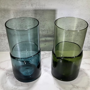 May include: Two cylindrical drinking glasses. One is teal, the other is olive green. Each glass has a dark base and a lighter, transparent upper section. The glasses are made of glass and are on a white marble surface.