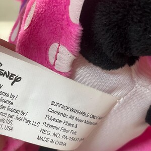 May include: A close-up of a Disney plush toy with a pink bow with white polka dots and black fur. A white tag with the Disney logo and text is visible. The tag reads "Surface Washable Only" and "Made in China."