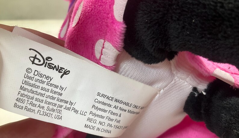 May include: A close-up of a Disney plush toy with a pink bow with white polka dots and black fur. A white tag with the Disney logo and text is visible. The tag reads "Surface Washable Only" and "Made in China."