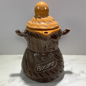 May include: A ceramic coffee pot with a lid and a handle. The pot is brown with a textured design and a tan lid. The word "Bocono" is written on the side of the pot.