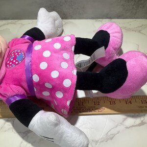 May include: A plush Minnie Mouse doll, lying on its back. The doll features a pink dress with white polka dots, black legs, pink shoes, and white gloves. A purple ribbon accents the dress. The doll has a tag attached.
