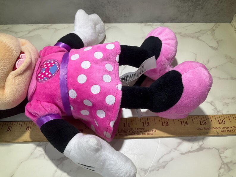 May include: A plush Minnie Mouse doll, lying on its back. The doll features a pink dress with white polka dots, black legs, pink shoes, and white gloves. A purple ribbon accents the dress. The doll has a tag attached.
