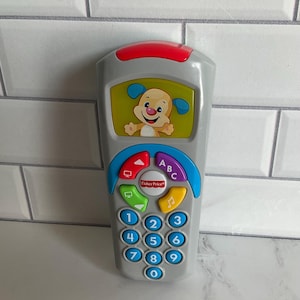 May include: A gray Fisher-Price toy remote control with a red top and a blue, red, and yellow button panel. The remote features a screen with a cartoon puppy and numbered buttons.