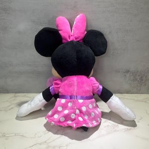 May include: A plush Minnie Mouse doll, viewed from the back, features a pink dress with white polka dots and a matching bow. The doll has black ears, black arms with white gloves, and a purple ribbon around the waist.