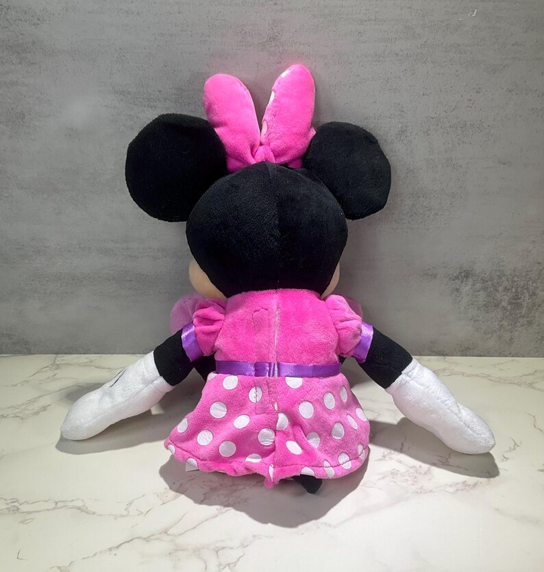 May include: A plush Minnie Mouse doll, viewed from the back, features a pink dress with white polka dots and a matching bow. The doll has black ears, black arms with white gloves, and a purple ribbon around the waist.