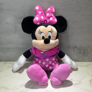 May include: A plush Minnie Mouse doll with black ears, a pink polka-dot bow, and a pink dress with white polka dots. The doll has a heart-shaped emblem on the chest and is sitting with arms and legs extended. The doll's face is beige with black eyes and a black nose.