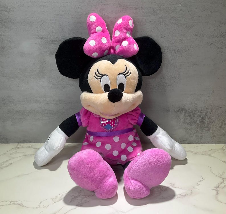 May include: A plush Minnie Mouse doll with black ears, a pink polka-dot bow, and a pink dress with white polka dots. The doll has a heart-shaped emblem on the chest and is sitting with arms and legs extended. The doll's face is beige with black eyes and a black nose.